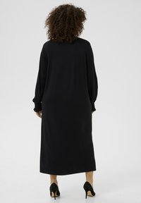 Vestido negro de manga larga hecho de tela suave, con puños fruncidos y una silueta recta, combinado con zapatos de tacón alto negros.
