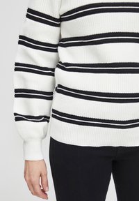 Pull en tricot blanc avec des rayures horizontales noires et blanches, poignets et ourlet côtelés. Associé à un pantalon de couleur sombre.