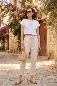 Kvinde iført solbriller, hvid crop-top t-shirt, beige bukser, der holder en lille flettet håndtaske på en brostensbelagt gade med blomster og stenhuse.
