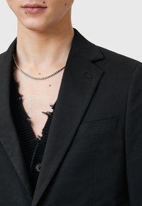 Svart blazer med slagkrage, en knappstängning och en framficka. Under finns en texturerad svart plagg med fransade kanter.