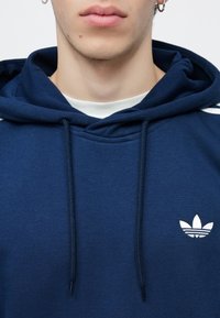 Marineblauwe hoodie van een katoenmengsel, met een voorkangoeroezak, een capuchon met trekkoord en een wit geborduurd logo op de borst.