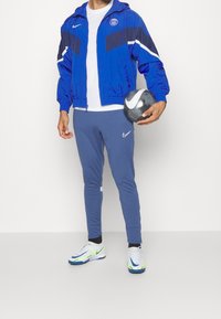 Person som bär en blå och marinblå sportjacka och byxor, håller en svart och vit Nike fotboll i sin högra hand.