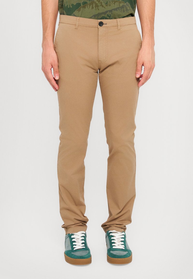 Chino in cotone beige con vestibilità slim, dotati di chiusura con un solo bottone, tasche laterali e polsini arrotolati. Abbinati a sneaker verdi e bianche.