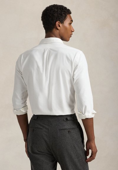 Hombre visto de espaldas que lleva una camisa blanca de manga larga con las mangas remangadas y pantalones entallados gris oscuro.