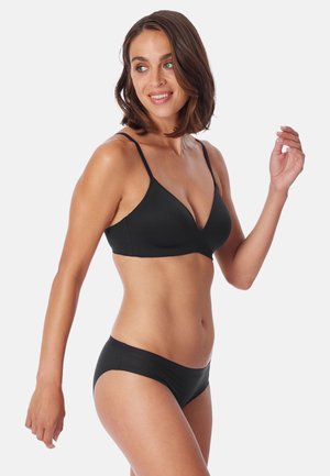 Kvinde iført en sort bh og matchende sort undertøj, der kigger til venstre med et let smil, mod en ensartet hvid baggrund.