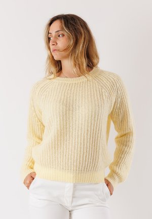 Donna in un maglione lavorato a maglia giallo chiaro e pantaloni bianchi, in piedi con le mani in tasca, che guarda di lato contro uno sfondo neutro.