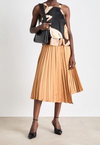 Haut noir sans manches avec accents à motifs ; jupe plissée beige ; escarpins noirs à bouts pointus avec brides à la cheville ; sac à main noir simple.