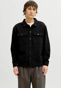 Młody mężczyzna o potarganych brązowych włosach ma na sobie czarną koszulę zapinaną na guziki z dwoma kieszeniami na piersi oraz luźne, szare spodnie na jednolitym tle.