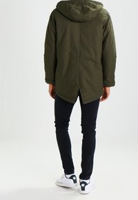 Olivgrön parka-jacka med huva, med en längre bakhem, sidofickor och strukturerad tyg. Kombineras med mörka jeans och vita skor.