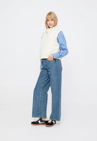 Bela flisasta brezrokavnik čez modro črtasto srajco, v kombinaciji z širokimi modrimi kavbojkami in črnimi teniskami. Preprosta in sproščena zasnova obleke.