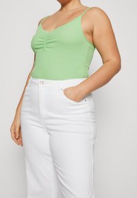 Top sin mangas verde con canalé y adornos de encaje, con detalle fruncido en el centro. Combinado con pantalones de mezclilla blancos de cintura alta, con el bolsillo delantero visible.
