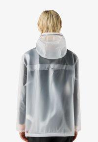 Person mit kurzen blonden Haaren, trägt eine durchscheinende graue Regenjacke mit Kapuze, von hinten vor einem schlichten Hintergrund gezeigt.