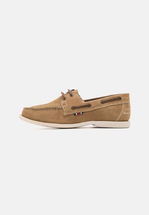 Chaussure bateau en daim beige avec semelle en caoutchouc blanche. Elle se distingue par des lacets marron, des détails cousus, et une petite accentuation multicolore sur le côté.