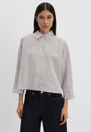 Vrouw draagt een wit overhemd met dunne zwarte verticale strepen en zwarte hoge taille broek, staande tegen een effen lichte achtergrond.