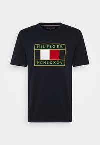 Černé tričko s krátkým rukávem, na hrudi s zeleným rámečkem a bílými a červenými vertikálními bloky, s textem "HILFIGER" ve zlaté barvě.