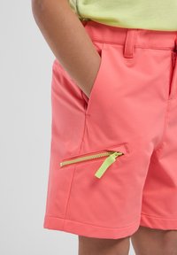 I pantaloni corti color corallo presentano una tasca con zip e un tira-zip di colore verde lime, realizzati in un materiale liscio e leggero.