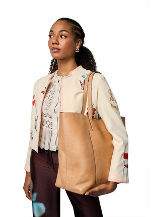 Lysebrun lædertaske med glat tekstur og minimalistisk design, med en sidelomme. Modellen bærer en broderet cremejakke over en blonder top.