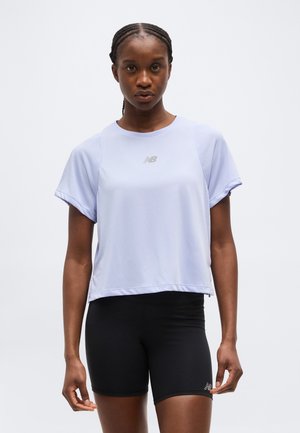 Atletisk kvinde med fletninger iført en lyse lilla kortærmet top og sorte tætsiddende shorts mod en ensartet baggrund.