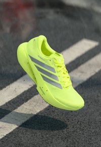 Πεπρωμένο κίτρινο παπούτσι για τρέξιμο με ασημί λεπτομέρειες, ελαφρύ σχέδιο, επάνω μέρος από δίχτυ και μαλακή σόλα, εμφανιζόμενο στον αέρα πάνω από ένα υφασμένο φόντο.