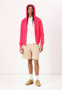 Roza zip-up hoodie s sprednjim žepom, v kombinaciji z belo majico in bež kratkimi hlačami. Model nosi bež čevlje in bele nogavice.