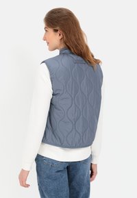 Gepolsterte ärmellose Weste in gedämpftem Blau, mit wellenförmiger Stickerei, Stehkragen und glatter Textur, kombiniert mit einem weißen Langarmhemd.