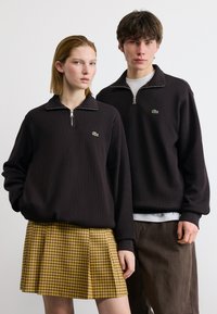 Νεαρή γυναίκα και άντρας φορούν ταιριαστά μαύρα φούτερ με φερμουάρ και λογότυπα Lacoste, στέκονται μπροστά σε απλό λευκό φόντο.