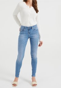 Bílý texturovaný pletený kardigan s knoflíkovým zapínáním, spárovaný s světle modrými skinny džíny. Modelka nosí béžové sand ály na otevřenou špičku.