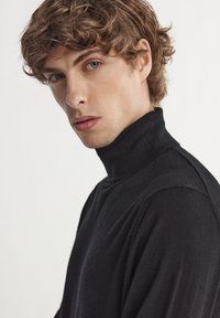 Schwarzer Rollkragenpullover aus weichem, strukturiertem Stoff, mit taillierter Passform und hohem Kragen. Der Stoff wirkt leicht und warm.