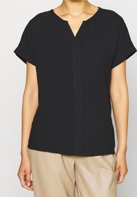 Blouse noire à manches courtes avec un col en V, en tissu doux, coupe ample et détails de couture frontale subtils. Associée à un pantalon beige clair.