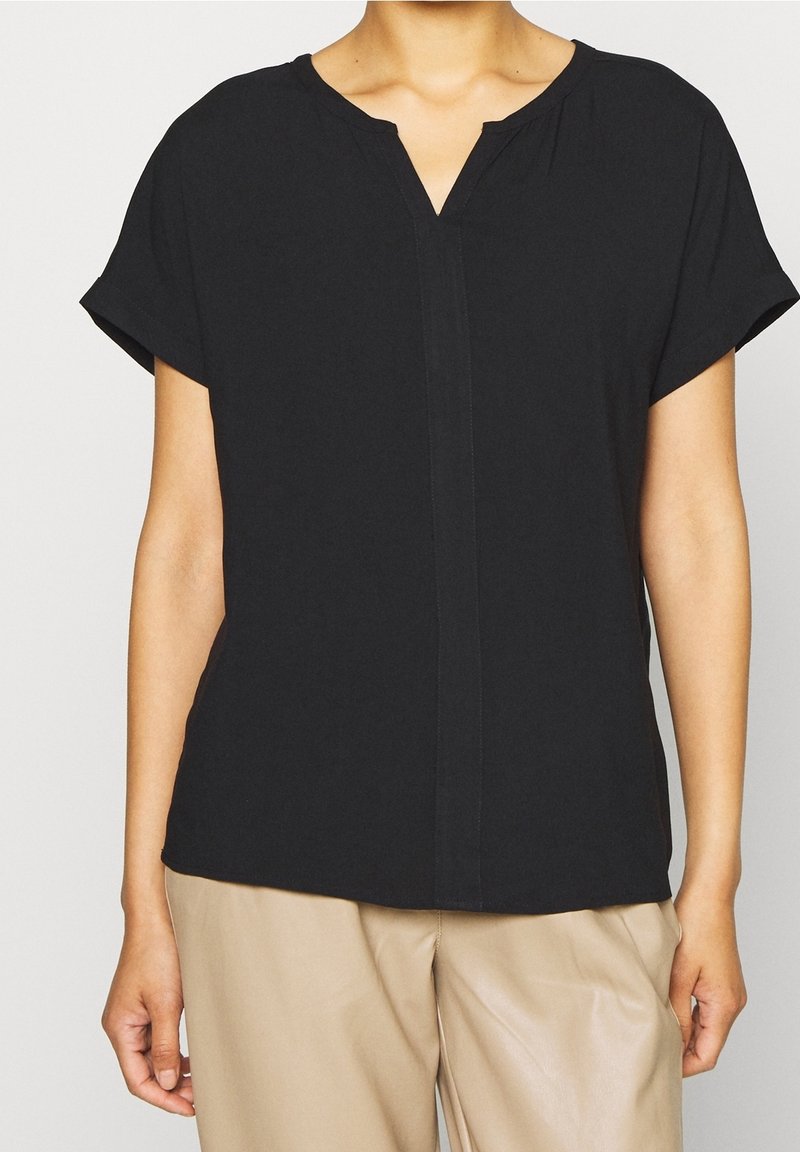 Blouse noire à manches courtes avec un col en V, en tissu doux, coupe ample et détails de couture frontale subtils. Associée à un pantalon beige clair.