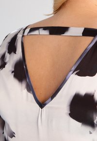 Blusa floreale bianco e nero con design a V sulla schiena, tessuto setoso e spalline scure a contrasto sulle spalle.