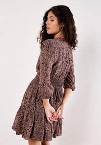 Frau mit lockigem Haar, die ein kastanienbraunes gemustertes Kleid mit 3/4-Ärmeln trägt und seitlich steht, mit Händen hinter dem Rücken verschränkt.