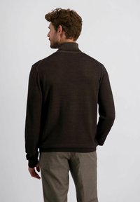Brauner Strickpullover mit einem hohen Kragen, der eine glatte Textur und gerippte Akzente aufweist. Kombiniert mit hellgrauen Hosen.