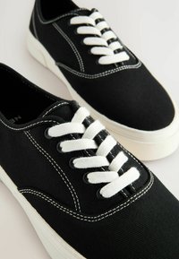 Scarpe da ginnastica in tela nera con lacci bianchi, cuciture a contrasto, punta arrotondata e suola in gomma. Design minimalista con silhouette classica.