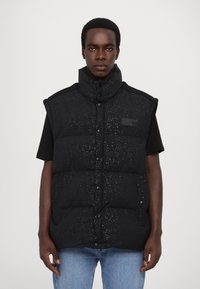 Black puffer vest with a high collar and snap buttons, featuring a subtle textured pattern and a rectangular patch on the left side.