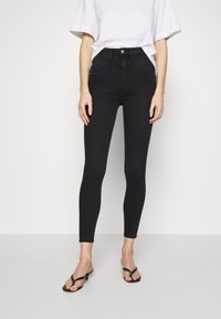 Czarne, obcisłe jeansy z wysokim stanem o gładkiej teksturze denimu, z przednimi kieszeniami i subtelnymi detalami szwów, zestawione z czarnymi klapkami na płaskiej podeszwie.