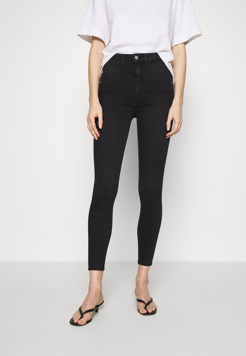 Czarne, obcisłe jeansy z wysokim stanem o gładkiej teksturze denimu, z przednimi kieszeniami i subtelnymi detalami szwów, zestawione z czarnymi klapkami na płaskiej podeszwie.