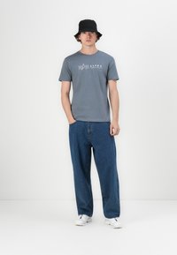 Pilkas medvilnės marškinėliai su baltu „Alpha Industries“ logotipu, derinami su laisvomis mėlynomis džinsinėmis kelnėmis ir juoda kibiro skrybėle. Pridedamos baltos sportbačiai.