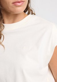 Cream-colored cotton T-shirt with a round neckline, short sleeves, and a subtle embossed logo on the left side. Smooth texture.