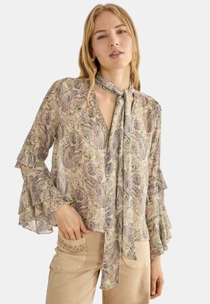 Joven mujer con una blusa de manga larga con estampado paisley, con mangas de volantes escalonados y un pañuelo anudado al cuello, combinada con pantalones beige con bolsillos delanteros.