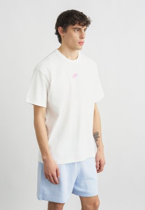 Jeune homme aux cheveux bouclés portant un t-shirt blanc Nike et un short bleu clair, debout devant un fond uni gris clair, tatouage visible sur le bras gauche.