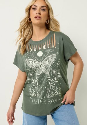 Lyshåret kvinde iført olivengrøn T-shirt med udskåret halsudskæring med hvid sommerfugl, månefaser, blomster, krystaller og teksten "Mystic Soul".