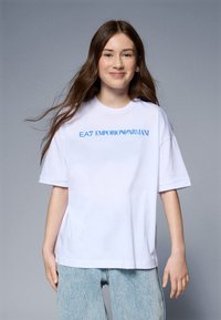 Bela prevelika bombažna majica s kratkimi rokavi, ki ima modri logotip "EA7 EMPORIO ARMANI". Ima gladko teksturo; kombinirana je s svetlo modrimi kavbojkami.