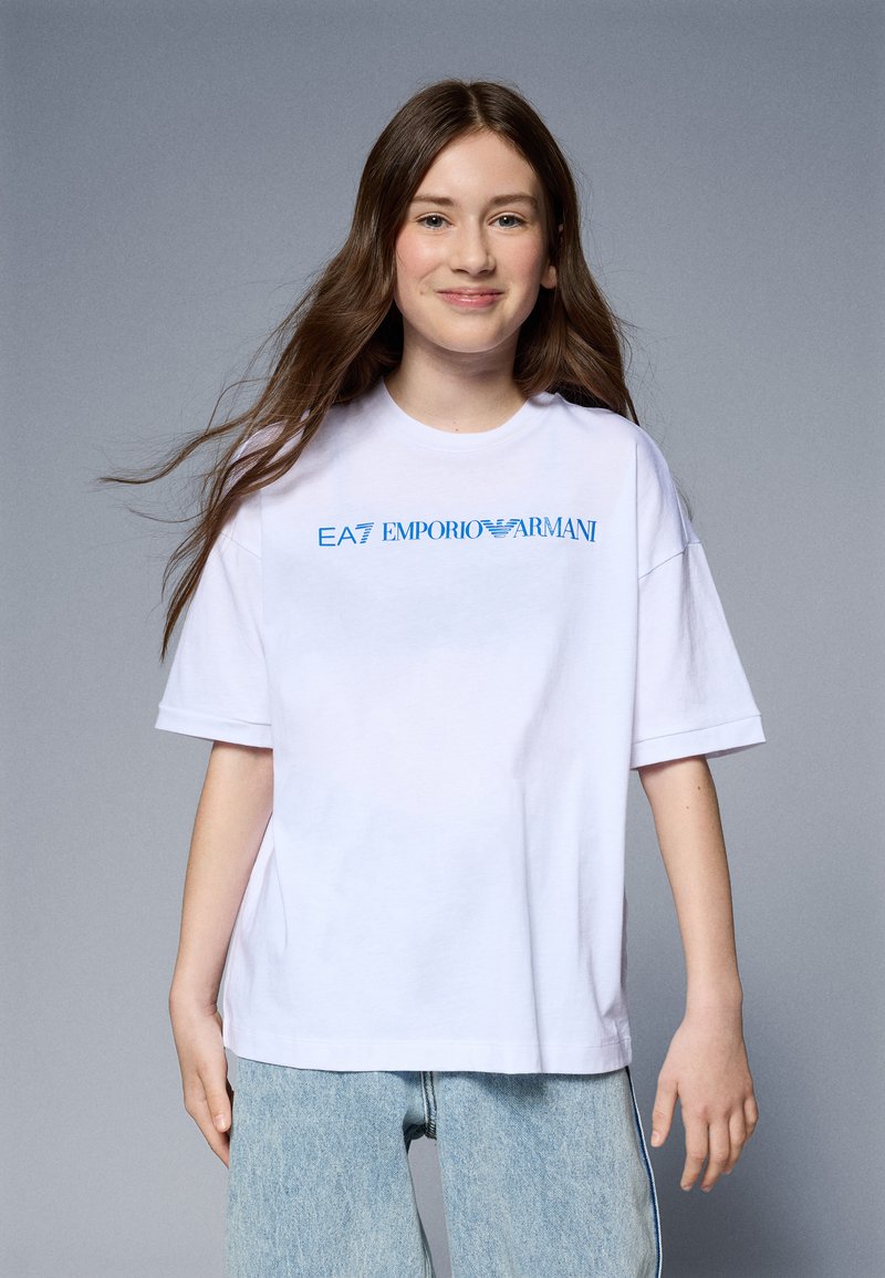 Bela prevelika bombažna majica s kratkimi rokavi, ki ima modri logotip "EA7 EMPORIO ARMANI". Ima gladko teksturo; kombinirana je s svetlo modrimi kavbojkami.