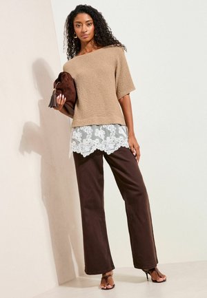 Žena nosi béžový pletený top cez bielu čipkovanú blúzku, tmavohnedé nohavice, čierne sandále na opätku, drží hnedé semišové púzdro s strapcami.