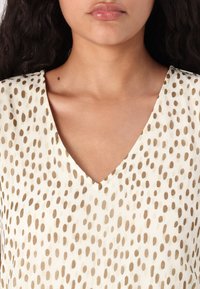 Femme portant un haut blanc à col en V avec des pois marron irréguliers, montré des épaules jusqu’à juste en dessous du cou.
