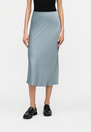 Frau trägt einen hellblauen Midirock und ein schwarzes ärmelloses Top, kombiniert mit glänzenden schwarzen Loafers, steht vor einem einfarbigen hellen Hintergrund.