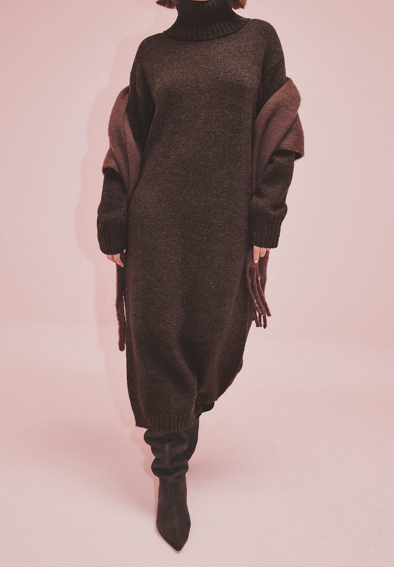 Personne portant une longue robe tricotée à col roulé marron foncé, un châle assorti drapé sur les épaules et des bottes noires décontractées sur un fond uni.