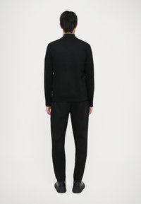Uomo in piedi di spalle, vestito con un completo tutto nero composto da una maglietta a maniche lunghe, pantaloni e scarpe nere, su uno sfondo bianco uniforme.