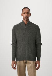 Dark green zip-up cardigan with ribbed texture, high collar, and long sleeves. Features silver zipper and finished hem. Worn over beige trousers.